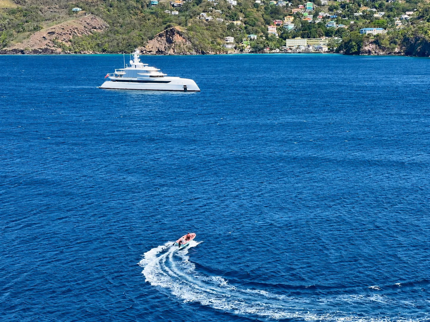 A motor yacht at anchor near a green coastline as a tender traces a curved wake across the bay.